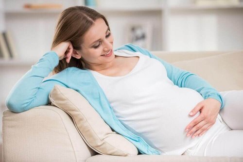 Eine schwangere Frau hält sich den Bauch und sitzt lächelnd auf einer Couch.