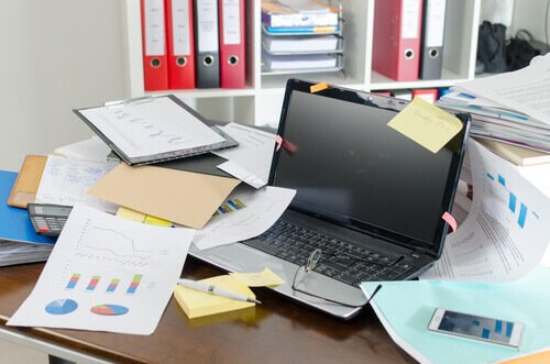Ein ungeordneter Schreibtisch voller Dokumente und einem Laptop führt zu Prokrastination.