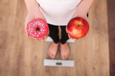 Frau steht auf der Waage mit Apfel und Donut in der Hand